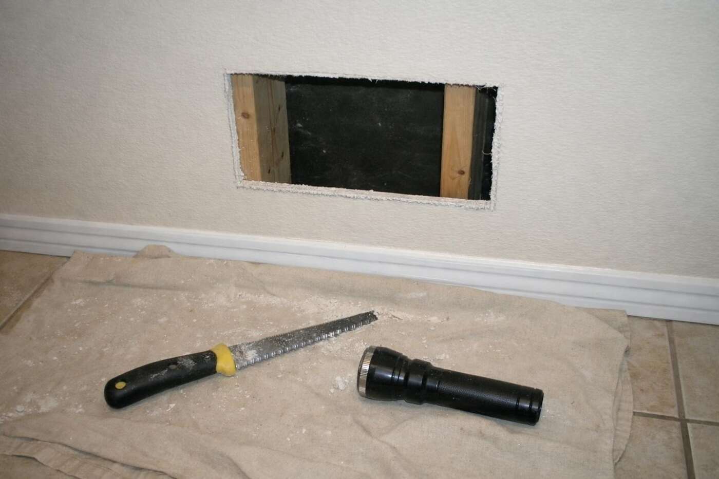 A small rectangular access opening cut into hallway drywall with a saw and light on the floor